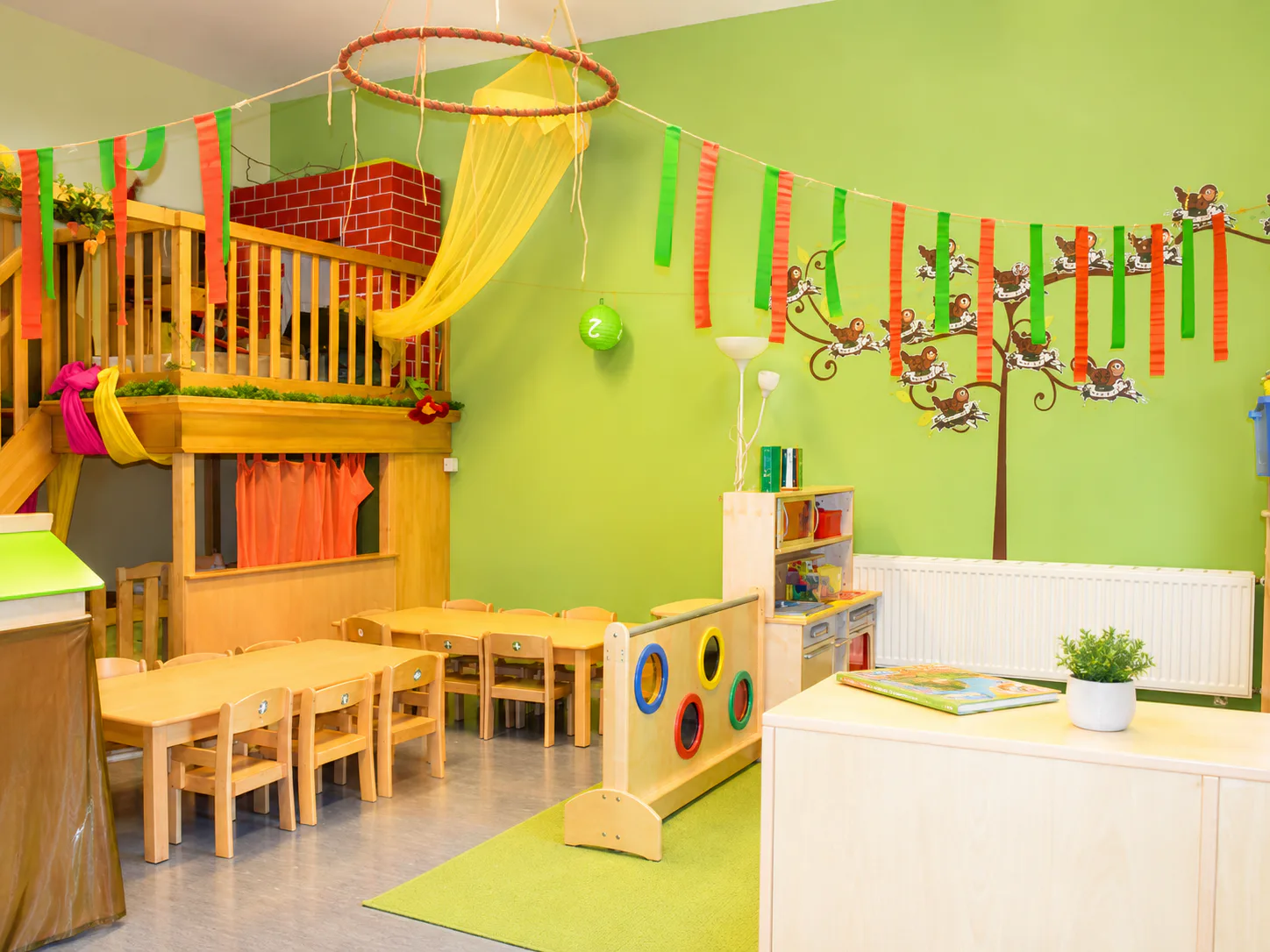 Grüner Gruppenraum mit Holzmöbeln, Spielpodest und Büchern